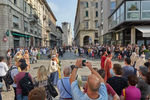 Como parade This urban photograph captures a lively scene in the city of Como, Italy during the afternoon in late summer. The image shows a parade taking place on a stone-paved street lined with historic buildings, with a crowd of people gathered on either side to watch the event. Many participants in the parade are dressed in period costumes, contributing to the festive atmosphere. In the background, the city’s medieval architecture is visible, including the prominent stone tower, Torre del Comune, which is a well-known landmark of Como. The bright sunlight and shadows indicate the time of day, while the attire of the people suggests that the season is summer. The photograph effectively documents a public celebration in urban Como, highlighting the vibrant culture and social life of this Italian city.